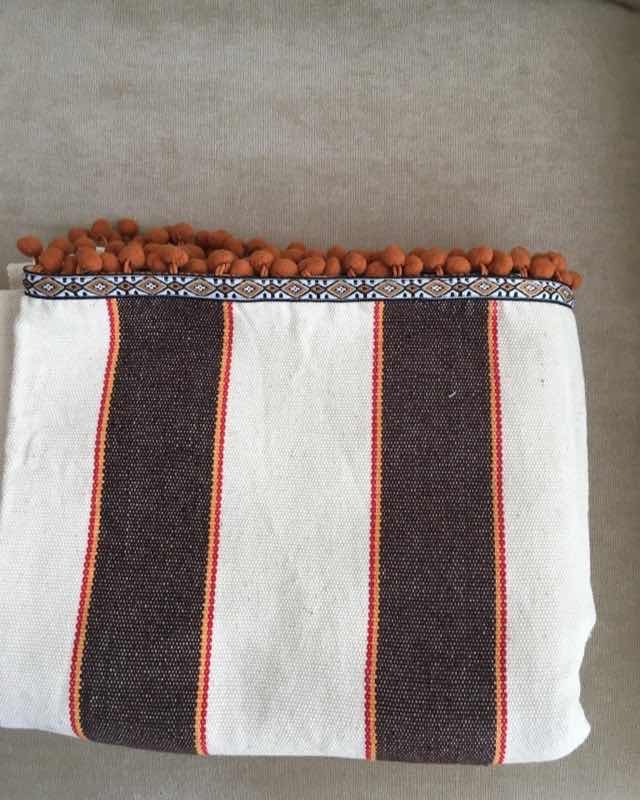 Mantel pequeño de algodón crudo con rayas café y naranja rematado con pompones